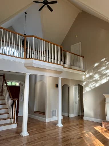 This is an image from Mendez Drywall & Painting LLC, located in Maryland. The image shows a spacious living area with high ceilings, white columns, beige walls, hardwood floors, and a staircase with white railings.