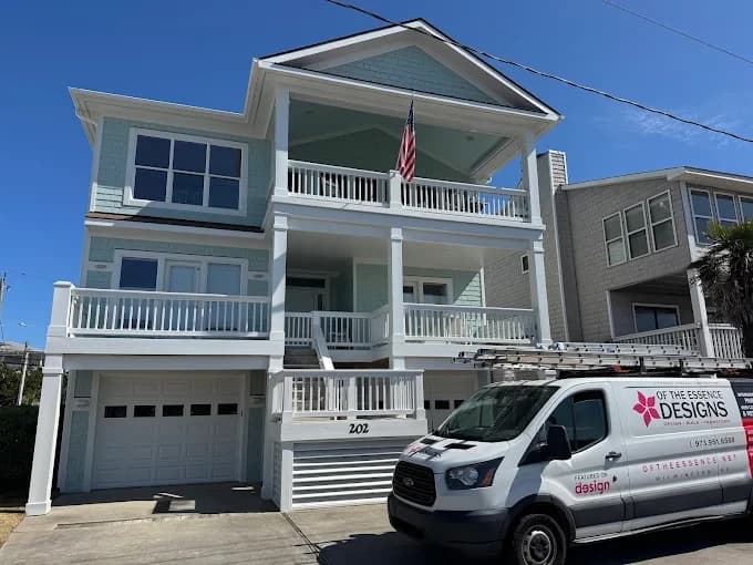 Exterior painting project by Of The Essence Design Build Paint in Wilmington, NC. The image shows a blue raised beach house with white railings, balcony, and garage.