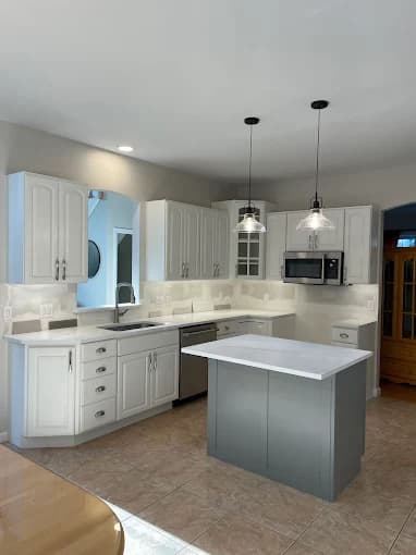 This is an image from JT’s Painting 518 LLC, located in New York. The image shows a freshly painted kitchen with white cabinets, light countertops, modern pendant lighting, and a contrasting gray island.