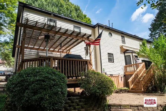 Exterior painting project by Garcia Paint Co in Greensboro, NC. The image shows a two-story house with light siding, dark trim, a wood deck, and pergola.