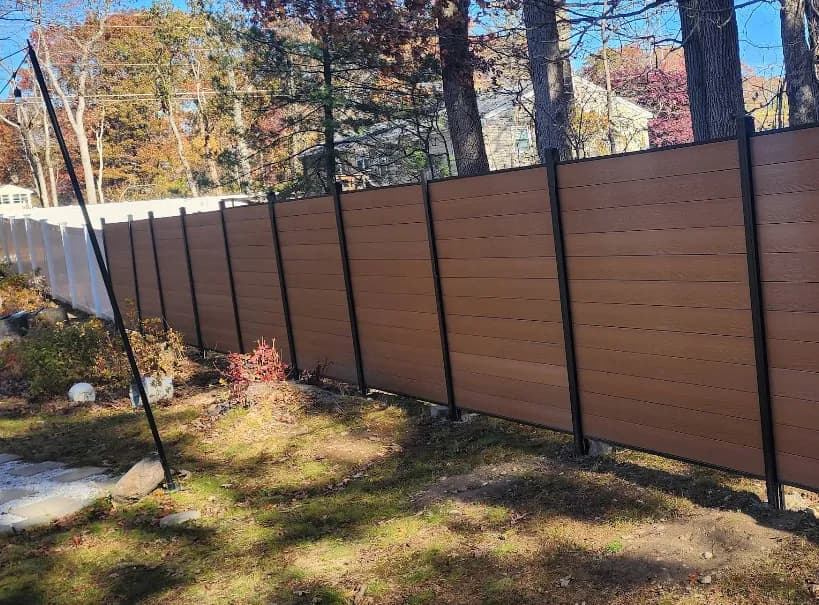This is an image from Nkomo Painting and Contracting, located in Massachusetts. The image shows a newly installed modern horizontal fence with brown composite panels and black metal posts along a wooded residential property.