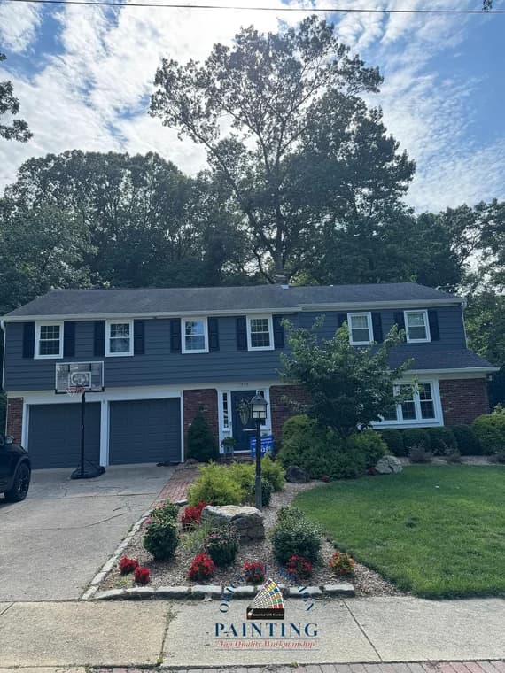 This is an image from Original Design Painting, featuring a large dark blue home with white trim and black shutters. The front yard includes vibrant red flowers and a clean paved walkway.