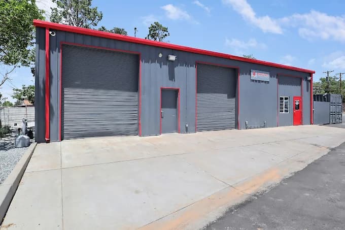 This is an image from Mike’s Quality Painting, located in New Mexico. The photo shows a freshly painted gray commercial building with bright red trim, large roll-up garage doors, and a clean concrete lot under a sunny blue sky.