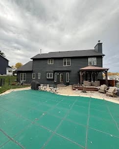 This is an image from Joseph Rivers Painting, located in Ohio. The photo shows a dark-painted two-story home with a closed green pool cover in the foreground, an outdoor seating area, and a wooden pergola on the right side.