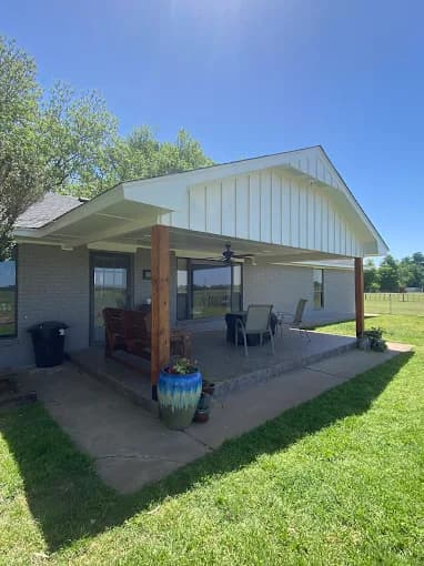This is an image from Fresh Coat Painters of Oklahoma City, located in Oklahoma. The photo shows a covered backyard patio with white board-and-batten siding, wooden support posts, and outdoor seating for dining and relaxation.