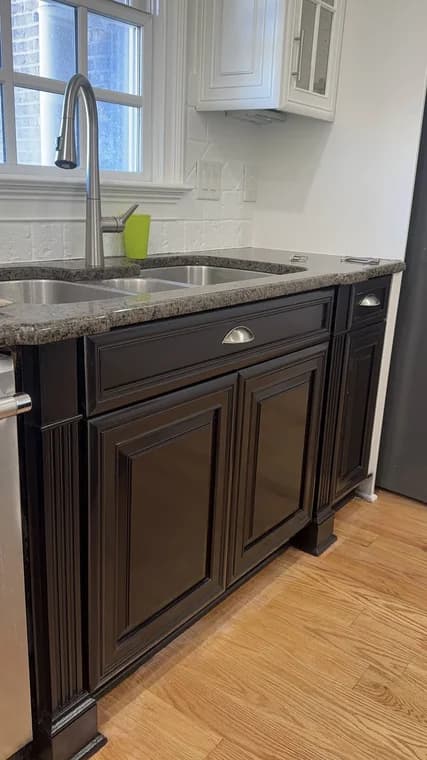 Cabinet painting project by Mario's Painting NC in Clayton, NC. The image shows dark kitchen cabinets, gray counters, a sink, white tile, and wood flooring.