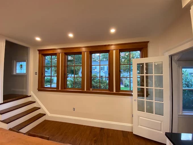 This is an image from Gonnella Painting & Restoration, located in Massachusetts. It features a renovated room with warm wood trim, large windows, recessed lighting, and cream-colored walls.