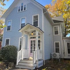 This is an image from Bates Brothers Painting LLC, located in New York. A freshly painted light blue two-story Victorian-style home with white trim, a covered front porch, and white railings surrounded by autumn foliage.