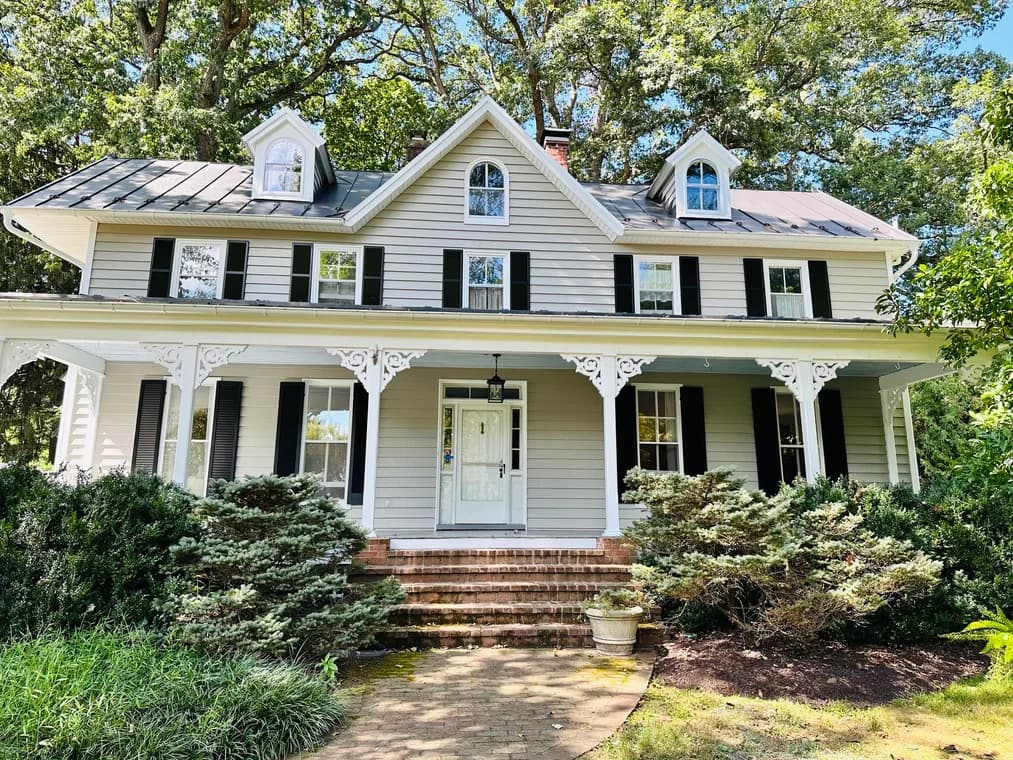 This is an image from Painting Contractors of MD, located in Maryland. The image shows a traditional two-story home with light siding, black shutters, a covered porch, and detailed white trim work.
