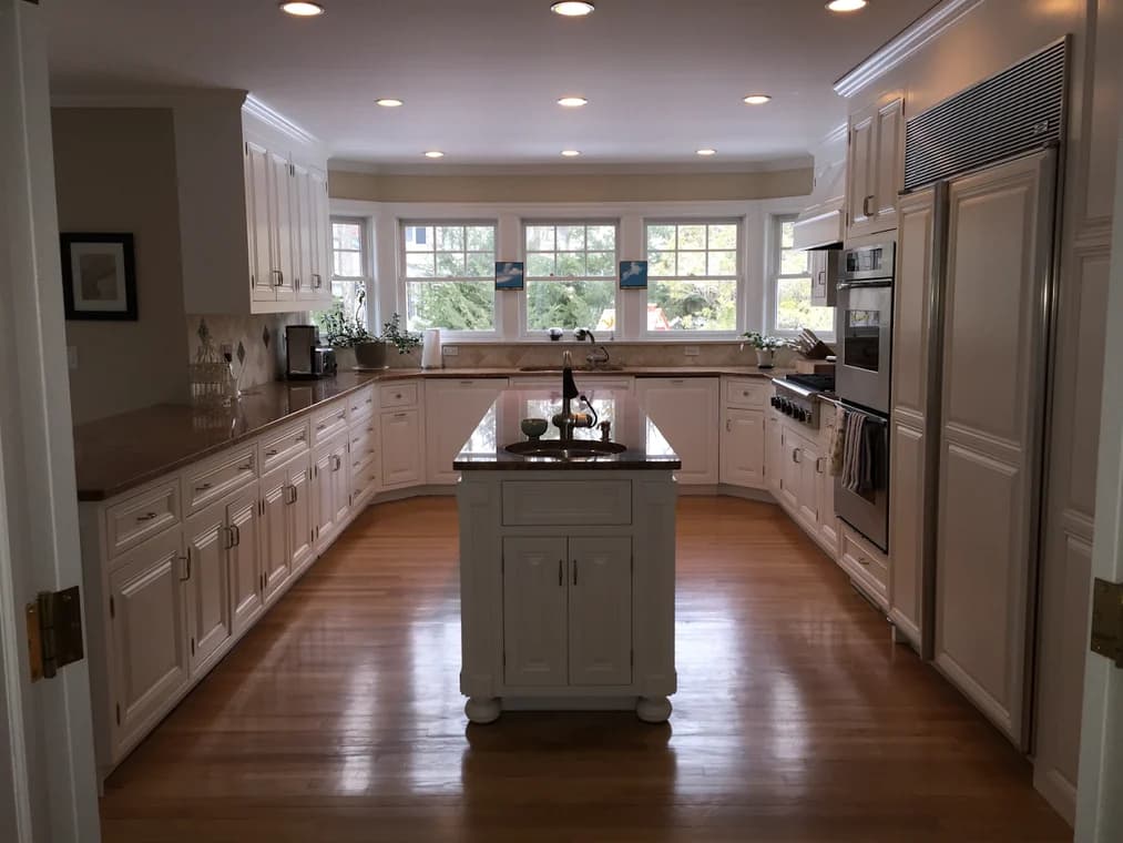 This is an image from J & S Painting Plus, located in New York. The interior photo features a spacious kitchen with white cabinetry, hardwood floors, a central island, and wide windows allowing natural light.