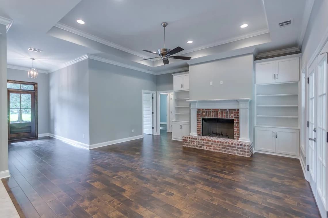 This is an image from Schmidt Painting, located in Massachusetts. It highlights an interior living room painted in a light gray tone by Schmidt Painting, with white trim, built-in shelving, and a red brick fireplace. The space features dark wood flooring and a ceiling fan.
