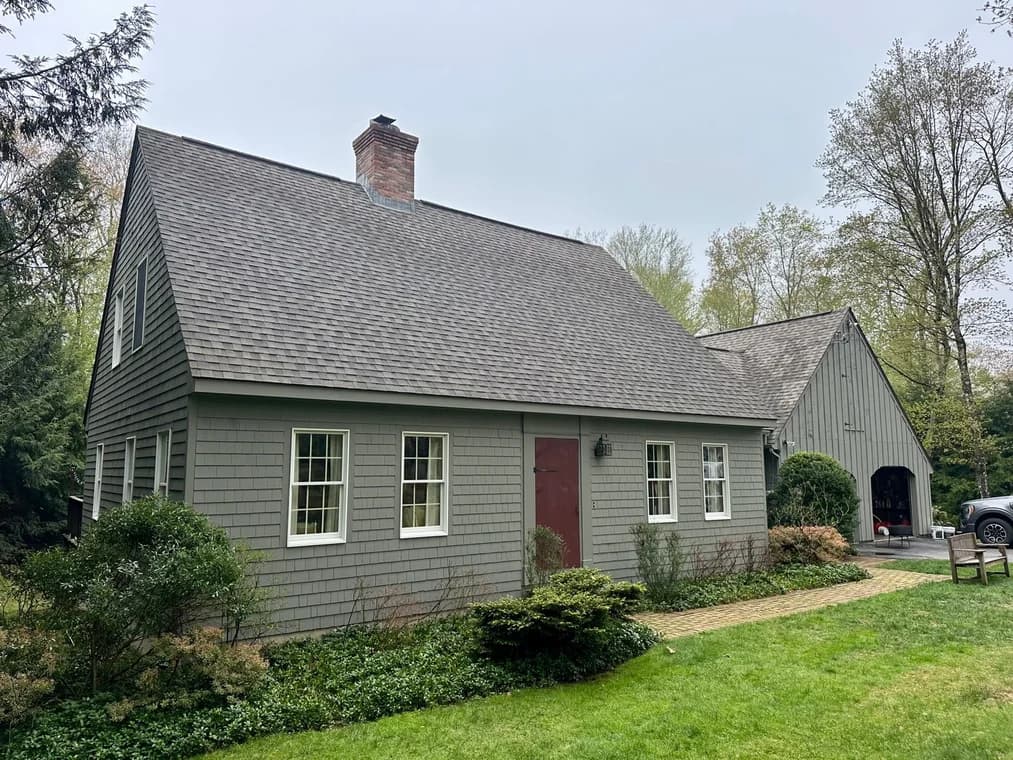 A gray-painted home with a steep roof, red door, and multiple white-trimmed windows, surrounded by green shrubs and trees.