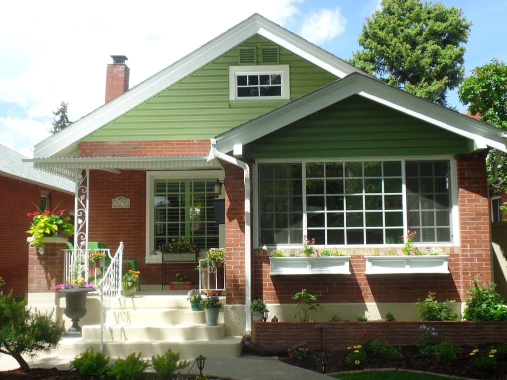This is an image from Pro Classic Painting company, located in Aurora, CO The image is a charming brick bungalow with freshly painted green gables and white trim, featuring decorative flower boxes and a well-maintained front garden, completed by Pro Classic Painting