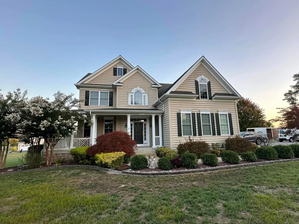 This is an image from DJ’s Painting & Powerwashing, located in Maryland. The image displays a tan two-story suburban home with white trim, black shutters, and landscaped shrubs across the front yard.
