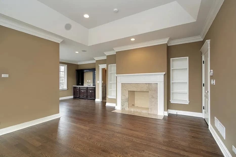 This is an image from Interior Finishing NYC, located in New York. The living room features freshly painted tan walls, white trim and crown molding, built-in shelving, and a large fireplace surrounded by dark hardwood floors.