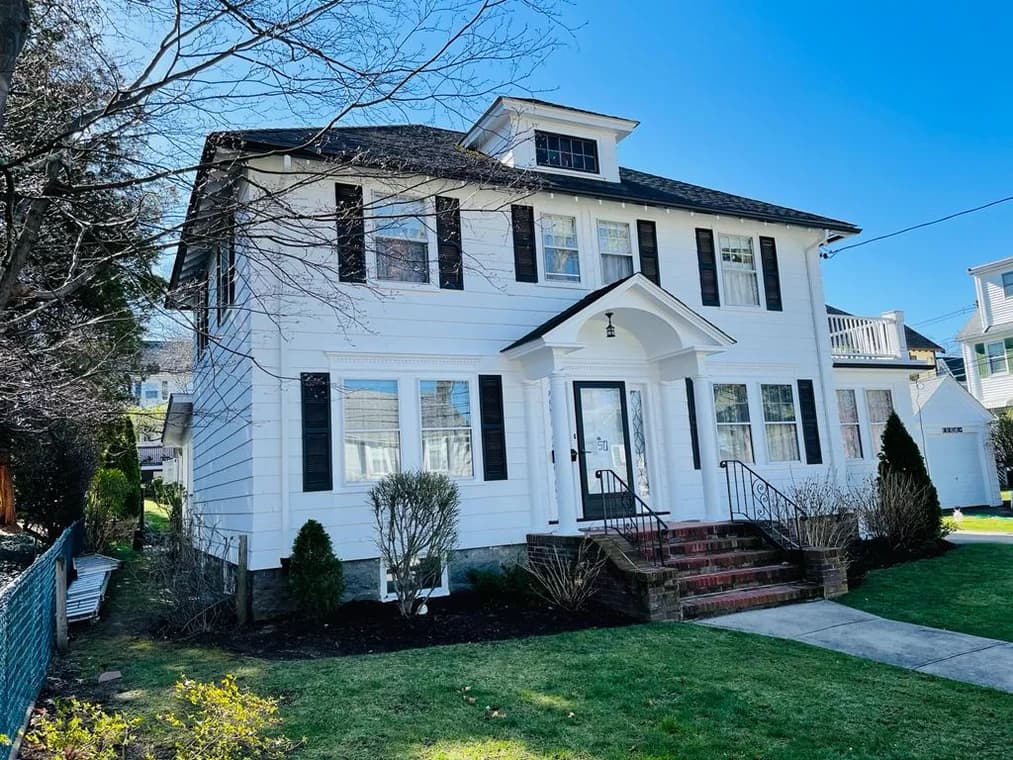 This is an exterior house painted by Rodrigo's Painting. The two-story colonial-style home is painted white with black shutters, a black roof, and a brick walkway leading to the front entrance.