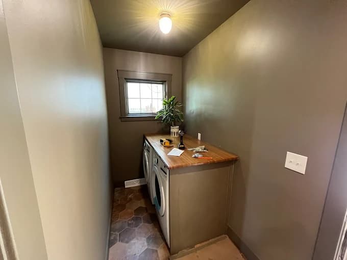 This is an image from BEE Pro Painting, located in New Jersey. The image shows a freshly painted laundry room with smooth gray walls, a dark ceiling, wood countertops, and updated trim around the window.