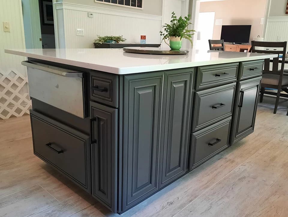 This is an image from Linda Loo Designs Cabinet Refinishing & Refacing, featuring a kitchen island with refinished gray cabinetry, black handles, and a white countertop. A green plant in a vase sits on top, adding a fresh touch.