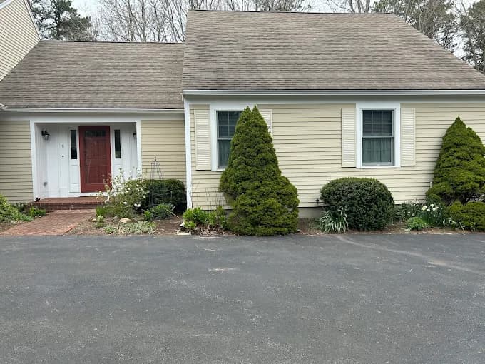 This is an image from Stewart Painting, located in Massachusetts. The photo shows a before view of a house with beige siding, green shutters, and a red front door prior to repainting.