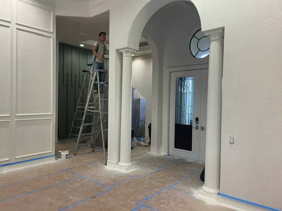 This is an image from Creative Colors And Design company, located in Naples, FL. The image is an interior painting in progress with painter on ladder near arched entryway and decorative columns in luxury home foyer by Creative Colors and Design