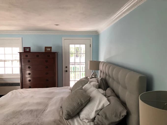 This is an image from CertaPro Painters of Southern Westchester NY, located in New York. The photo shows a bedroom with light blue painted walls, white crown molding, a tufted gray headboard, and natural light coming through large windows and a glass door.