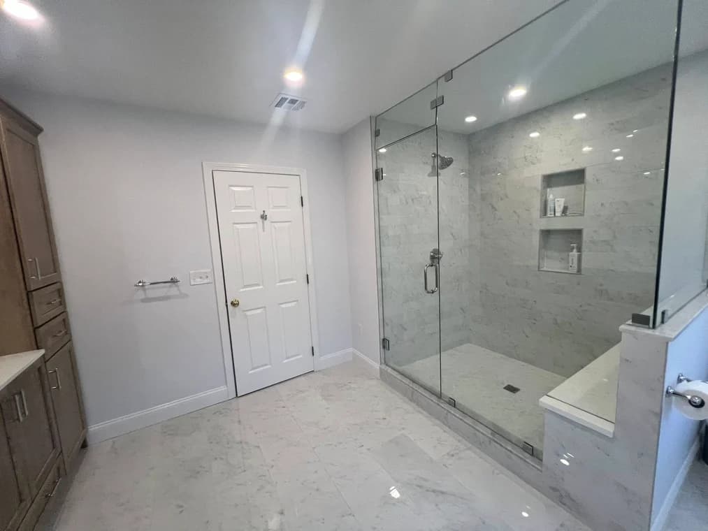 This is an image from Powell Painting & Home Services, located in Massachusetts. The photo shows a newly painted modern bathroom with light gray walls, a glass walk-in shower, and polished marble tile flooring.