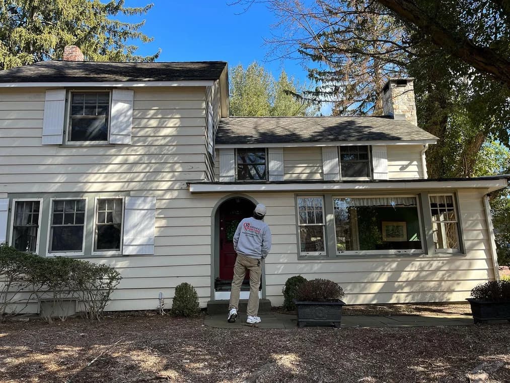 Exterior of a two-story cream-colored home with white shutters, where a painter in Romeo Painting Plus attire is inspecting the siding before work begins.