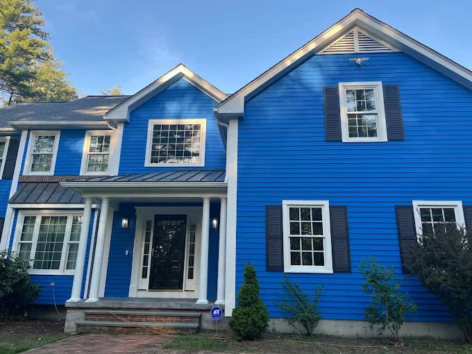 This is an image from Aurora Exterior Painting Inc., located in Massachusetts. The photo shows a large two-story home with bright blue siding, white trim, and dark shutters, highlighting a bold and refreshed exterior finish.